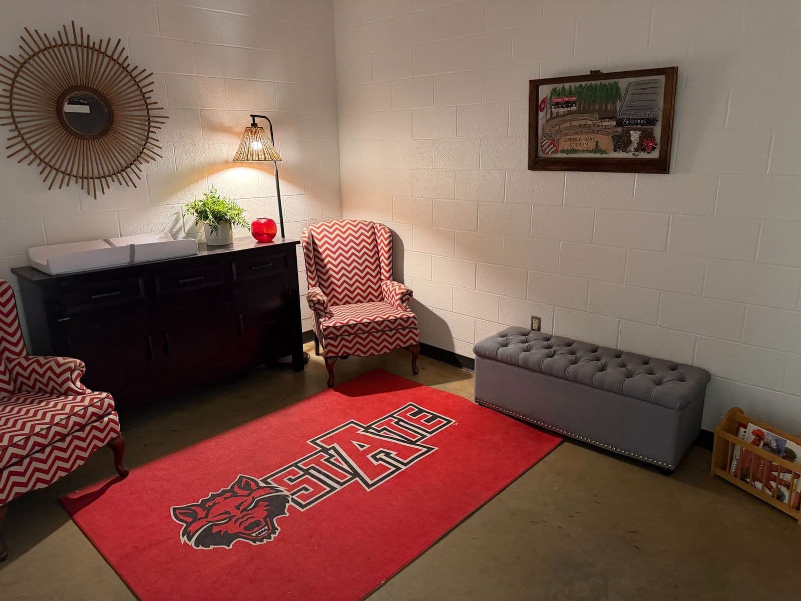 The lactation room at Centennial Bank Stadium: two red-and-white chevron armchairs, a changing table on a wooden cabinet, a STATE rug with the Red Wolves logo, and a book rack for children