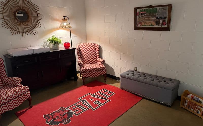 The lactation room at Centennial Bank Stadium: two red-and-white chevron armchairs, a changing table on a wooden cabinet, a STATE rug with the Red Wolves logo, and a book rack for children