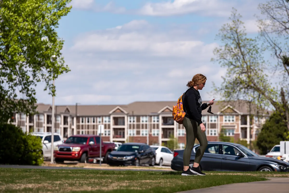 An A-State student walking across campus with the student apartments behind